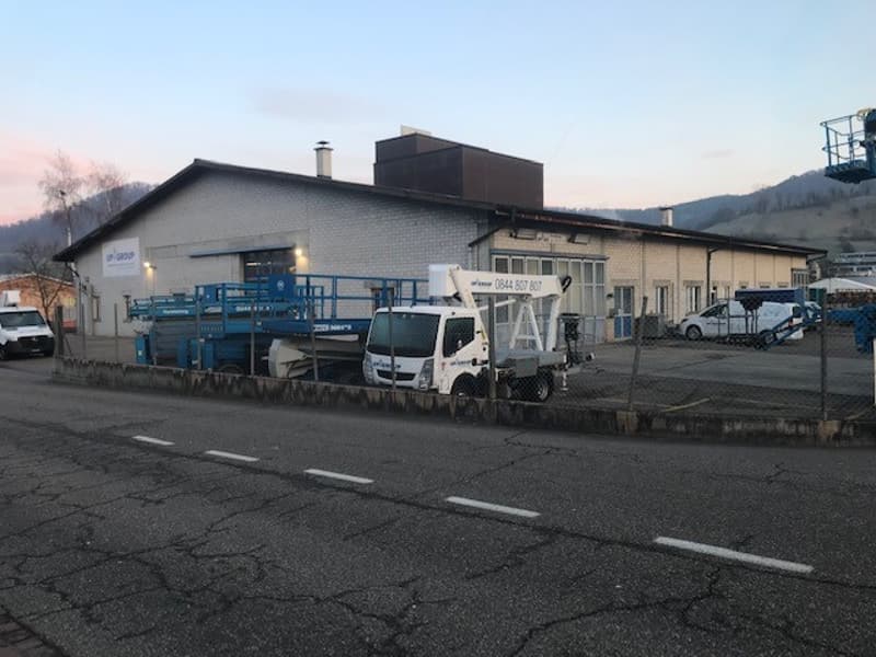 Industrial building with large entrance, blue lift, several parked vehicles, fence