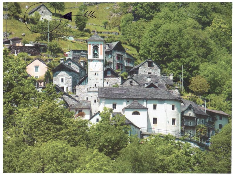 A picturesque village nestled in a lush, green valley. The buildings are made of stone and have traditional alpine architecture, with a church tower and several multi-story structures. The village appears to be built on a hillside, with some buildings hav