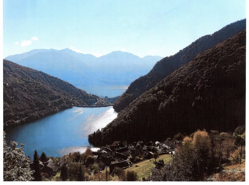 The image shows a scenic mountain landscape with a lake in the valley below. The mountains have a rugged, forested appearance, and the lake appears calm and reflective. There is a small village or town visible in the foreground, nestled among the hills. T