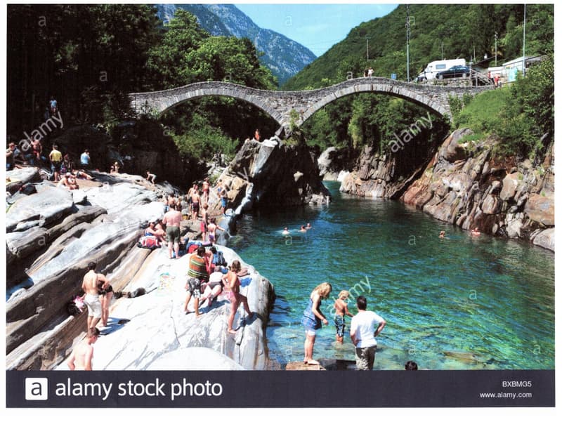 A scenic image of a stone arch bridge over a clear blue river surrounded by lush green mountains. Many people are swimming and relaxing on the rocks around the river.