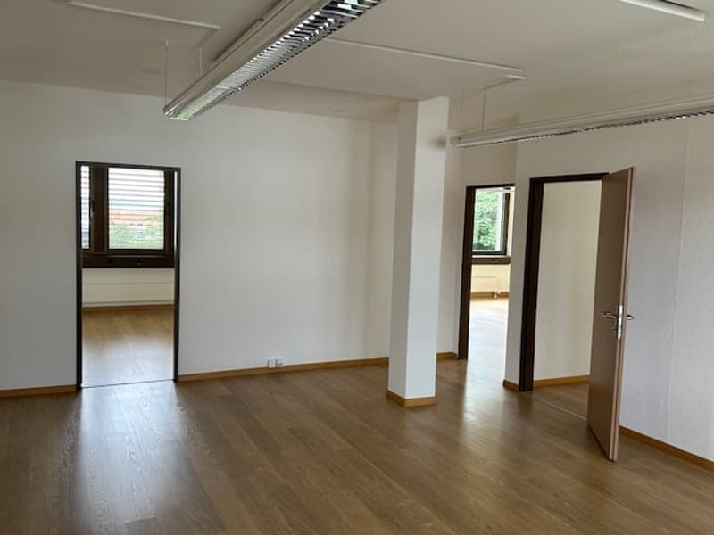 The image shows a large, open office space with white walls, hardwood floors, and large windows. The room has a high ceiling with overhead lighting fixtures. There are two doors visible, leading to other rooms or areas.