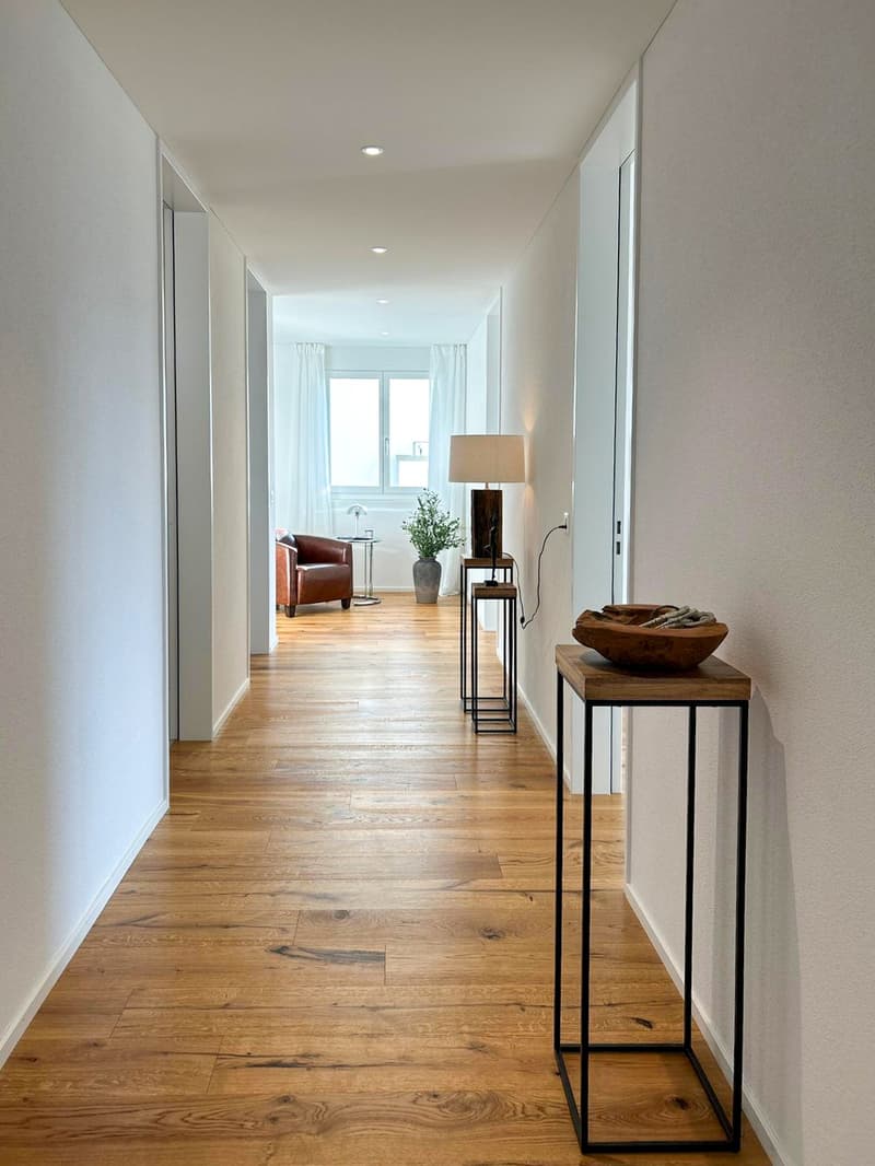 Wooden floor, white walls, small table with lamp, table with plant, couch, open window, hallway
