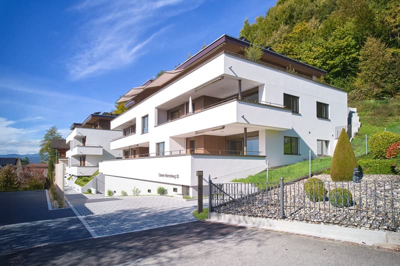 A modern, multi-story building with white exterior, multiple balconies, a driveway with gravel, and a hill with vegetation in the background.