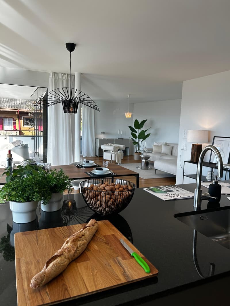modern kitchen, dining table, 2 lamps, couch, shelves, potted plants, black countertop, modern sink with faucet, wooden cutting board, green knife, bread on the cutting board