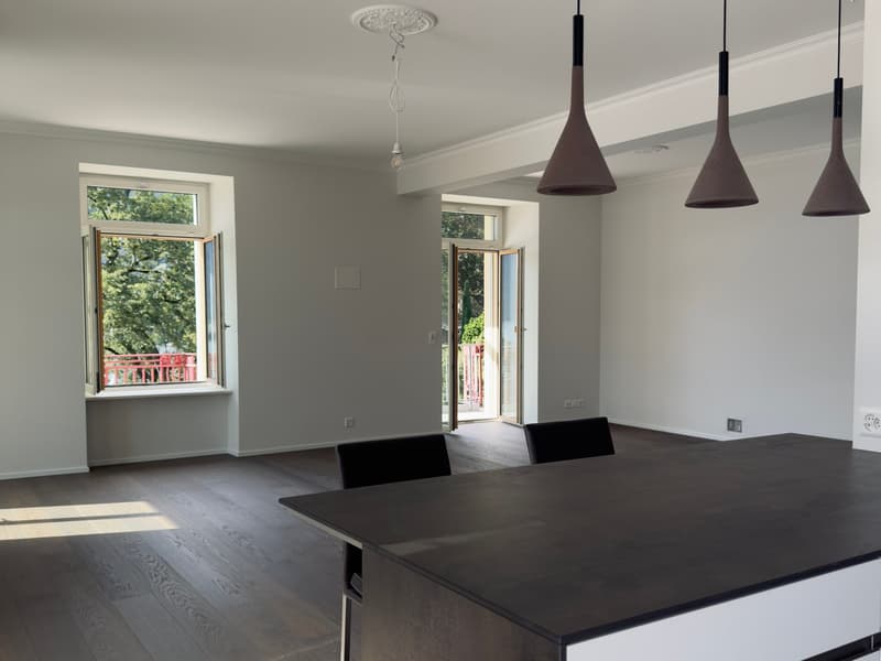 Empty room, white walls, black ceiling lights, hardwood floor, balcony with a view of the trees