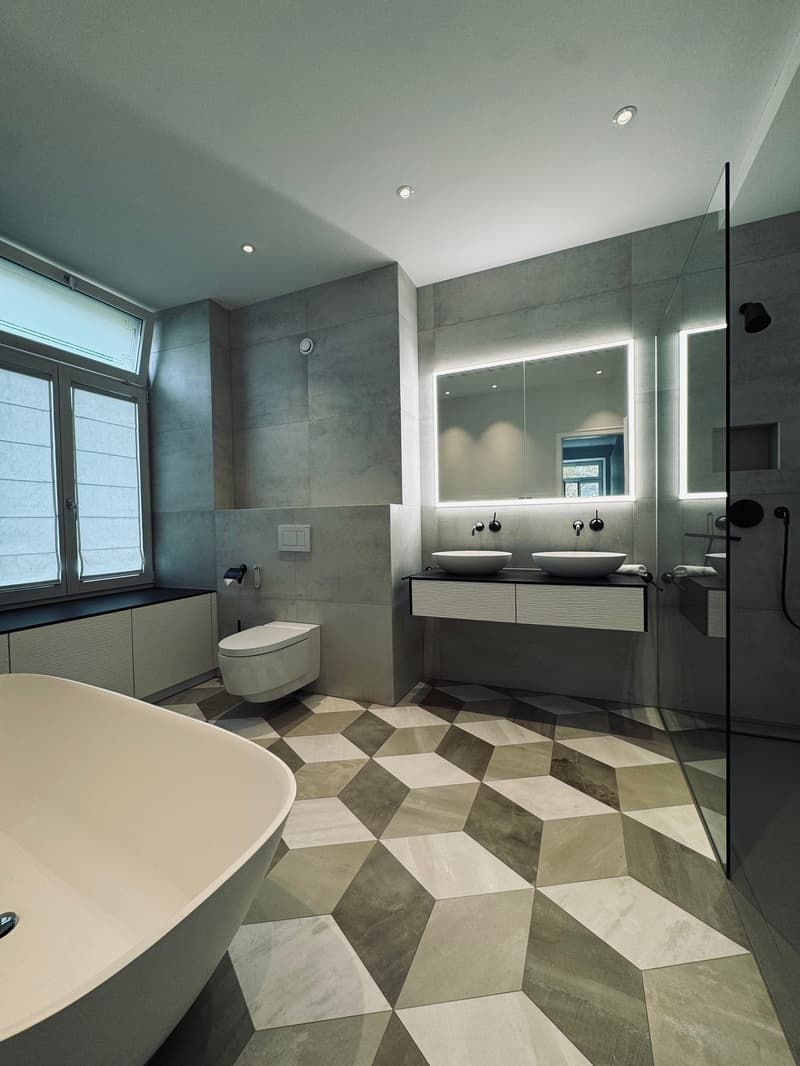 White oval bathtub, double washbasin, separate toilet, modern design, grey floor tiles, mirror, recessed lighting, toilet paper holder, towel holder
