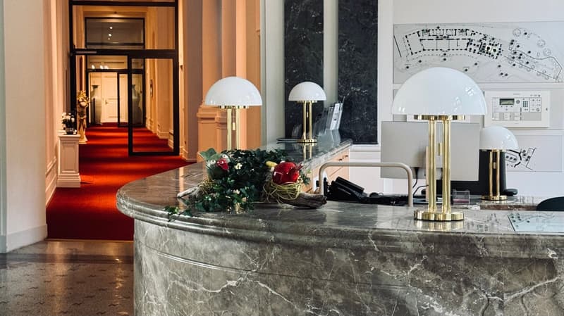 Luxurious hotel lobby with marble countertop, brass lamps, and a red carpet leading to the entrance. The lobby features a fireplace and various decorative elements such as flowers and artwork on the walls.