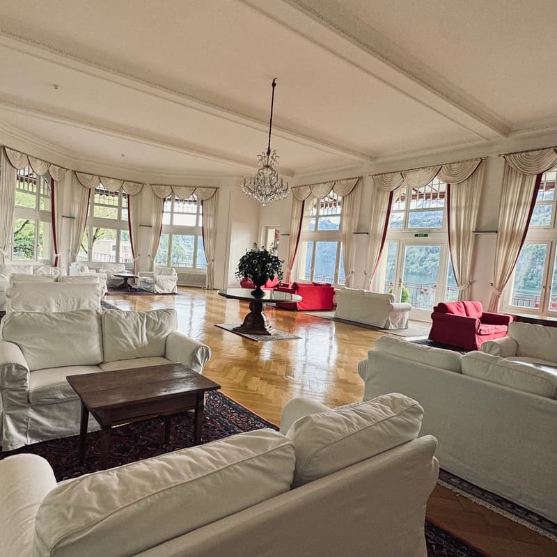 large living room with white and red couches, wooden table with plant, wooden floor, large windows with curtains, balcony