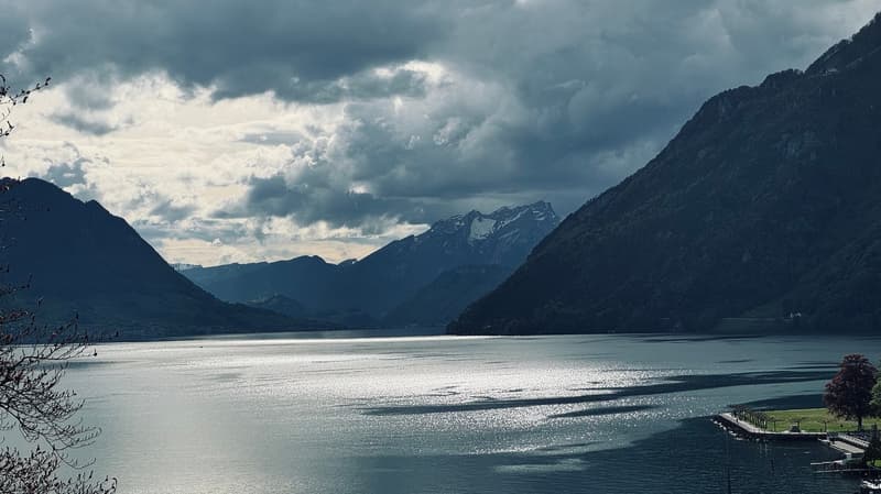 The image shows a scenic view of a mountainous landscape with a large lake in the foreground. The lake is surrounded by steep, snow-capped mountains and the sky is filled with dramatic, stormy clouds. The landscape has a moody, atmospheric feel with the d