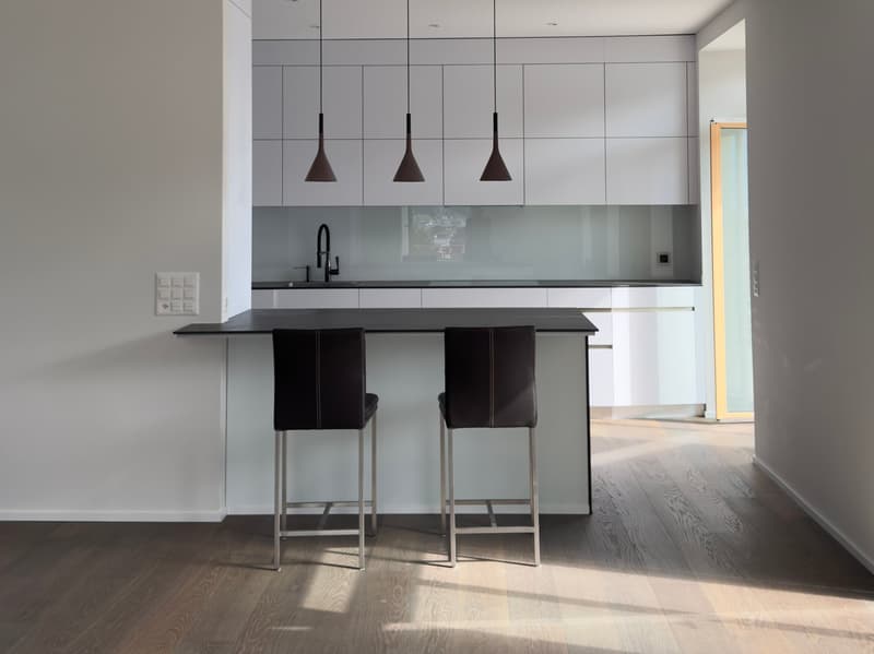Modern kitchen with white cabinets, black countertop, island with bar stools, glass backsplash, pendant lights, wooden floors, and a sink with faucet.