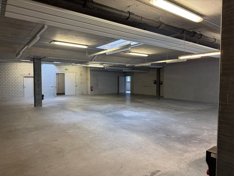 Empty warehouse room, concrete floor, brick wall, exposed beams, ceiling lights, emergency lights