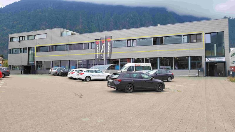 A multi-story commercial building with a parking lot in front. The building has large windows and signage for various businesses, including Facility Services and Eco Cristina. The building is surrounded by mountains in the background.