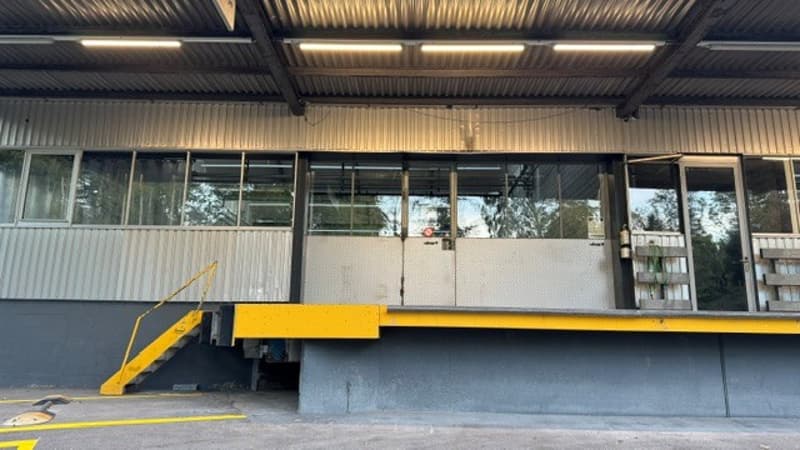 Industrial-style building with a large garage entrance, yellow loading ramp, and multiple windows. The structure features a metal roof and walls, with a concrete base and a yellow staircase leading up to the entrance.