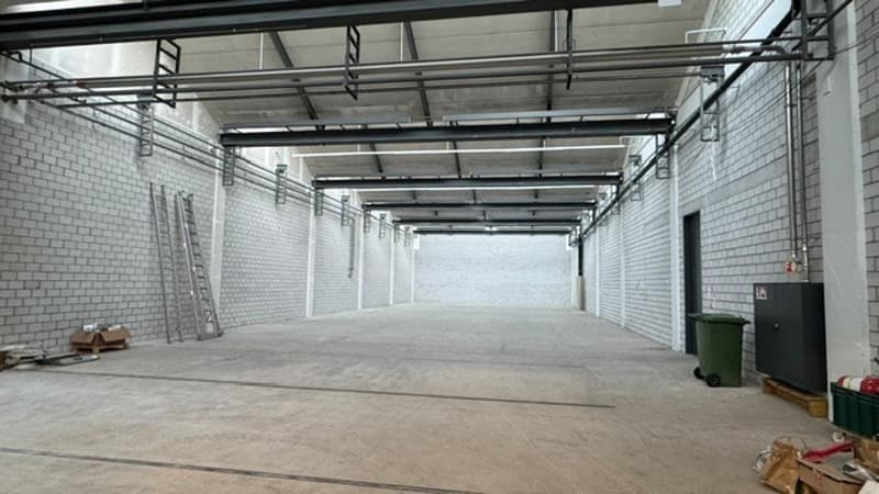 Empty industrial-style warehouse space with exposed brick walls, metal ceiling beams, and concrete floor. Minimal furnishings include a ladder, a few storage boxes, and a green waste bin.