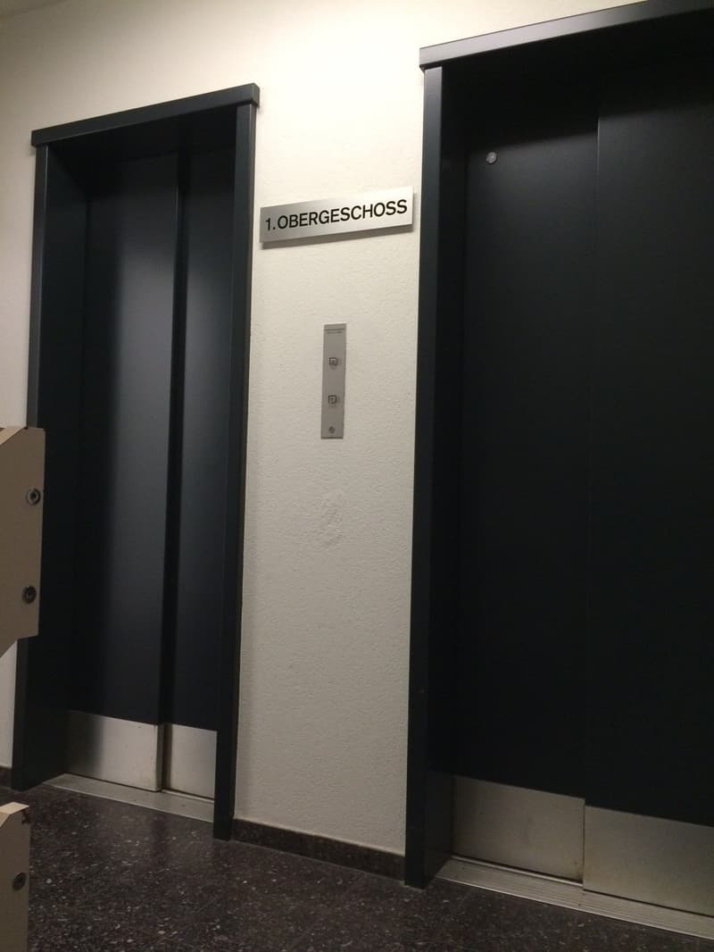 The image shows the entrance to a building, with two black elevator doors and a sign that says '1. Obergeschoss', which means 'First Floor' in German.