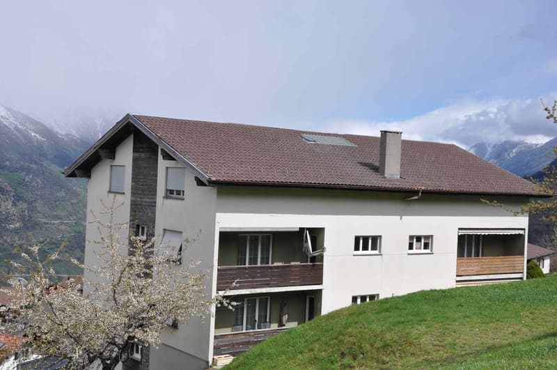 Three story building, brown roof, white walls, multiple windows, balcony, located on a slope with grassy terrain and mountain view