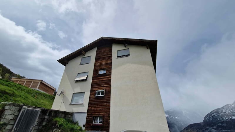 White and brown exterior, multiple windows, satellite dish on wall, mountain view, another building behind the hill