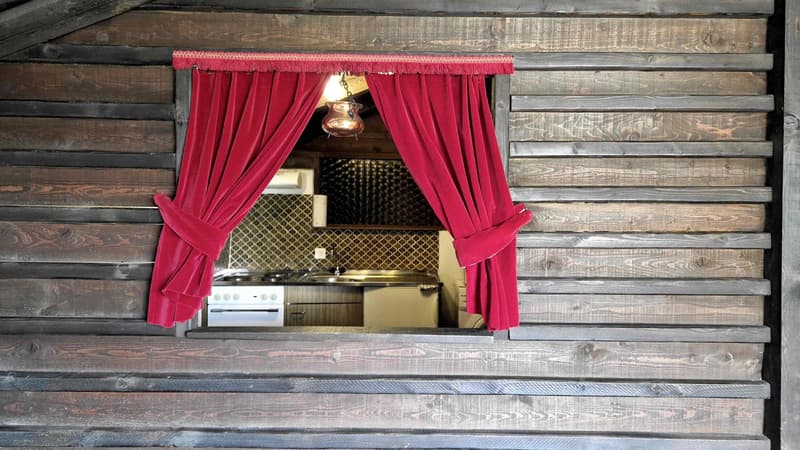 The kitchen has a window with red curtains, a copper lamp, a tiled backsplash, an oven, a sink with a faucet, a refrigerator, and wooden cabinets.