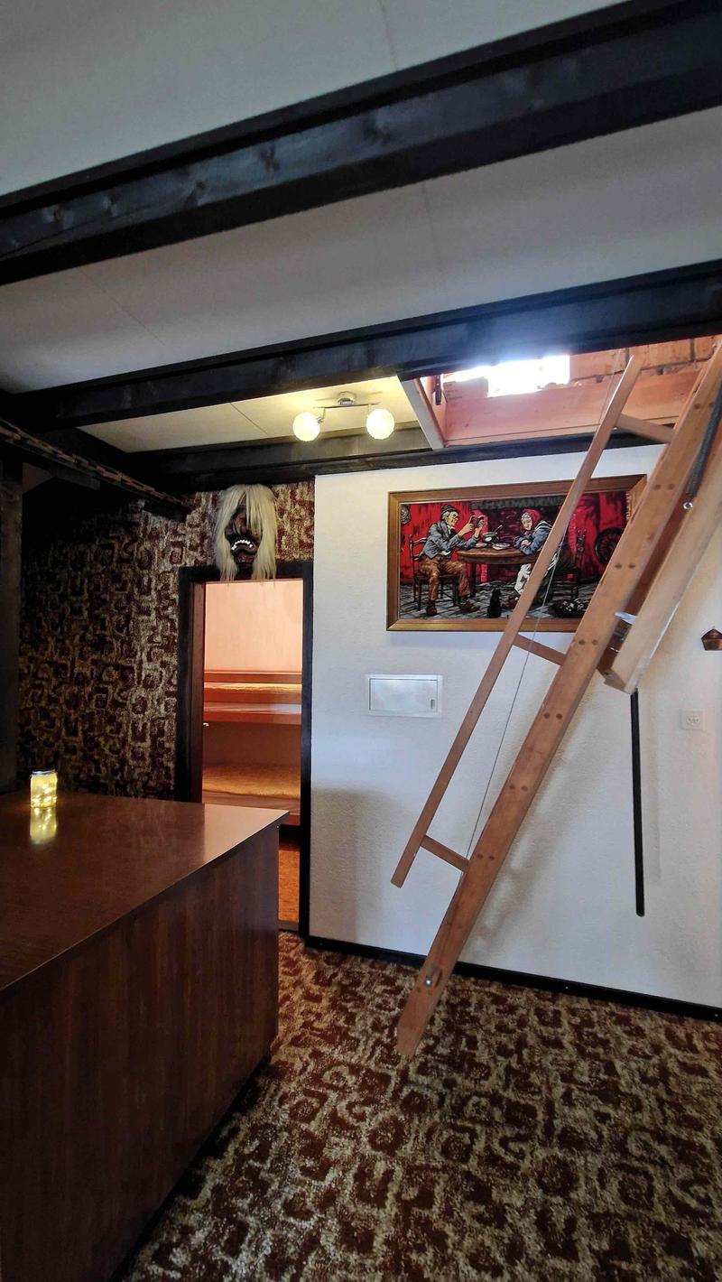 A room with carpet flooring, a wooden ladder attached to the wall, a desk with a candle on it, a framed picture, a small door leading to another room, and a wooden mannequin on the wall.