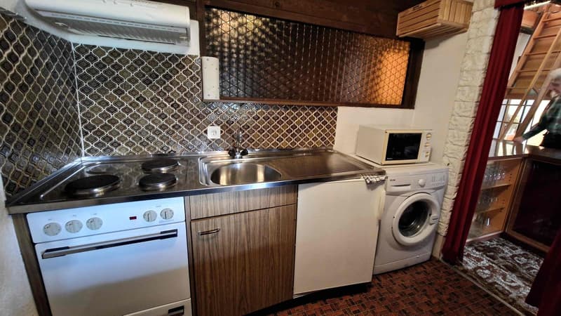 Compact kitchen with a stainless steel countertop, an electric stove, sink, and wooden cabinets. Appliances include a white microwave and a white washing machine. The wall has a tile design, and an open doorway to another room is visible on the right.