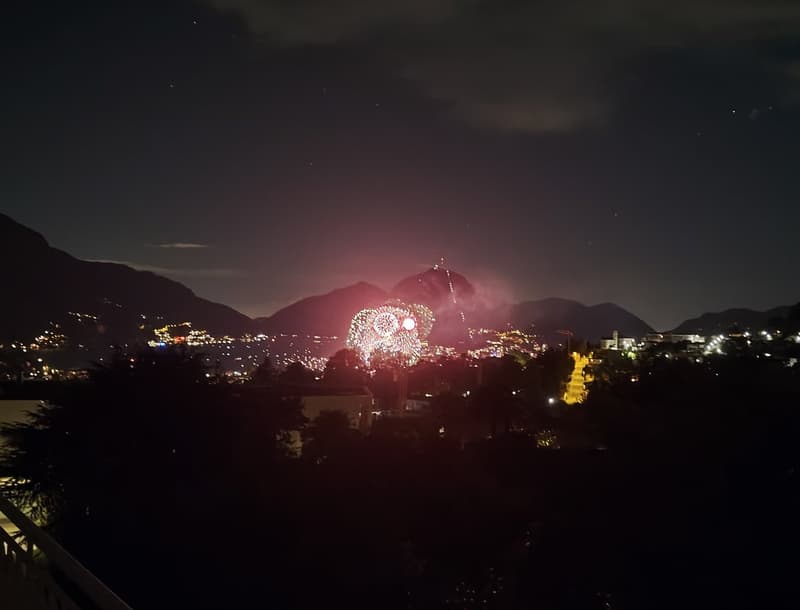 Night view of a city with fireworks, buildings, and mountains in the background.