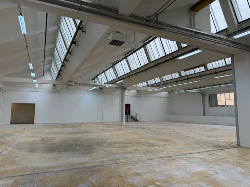 Spacious interior of a building with metal beams and skylights, concrete floor, two doors, one on the left and one on the right, a staircase in the center.