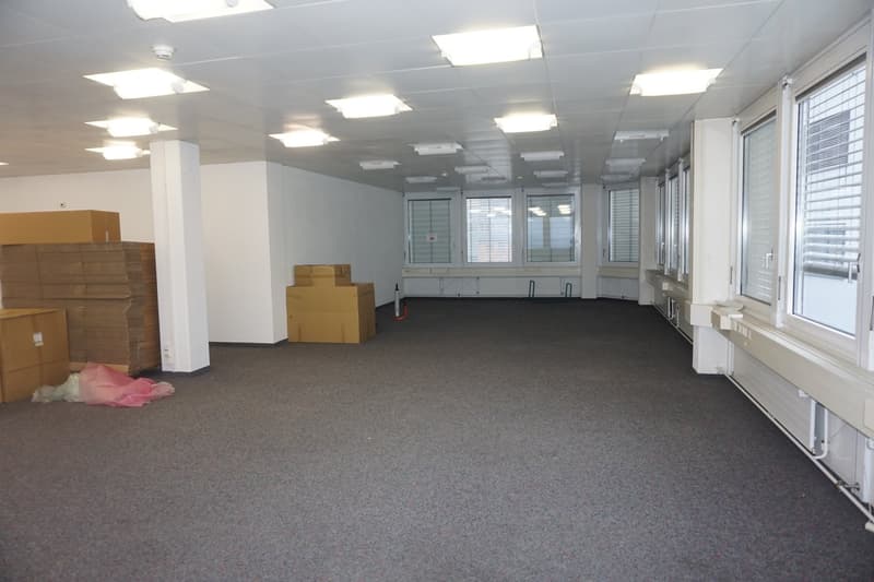 Large open room, carpet flooring, numerous large windows, pillars in the center, storage boxes present