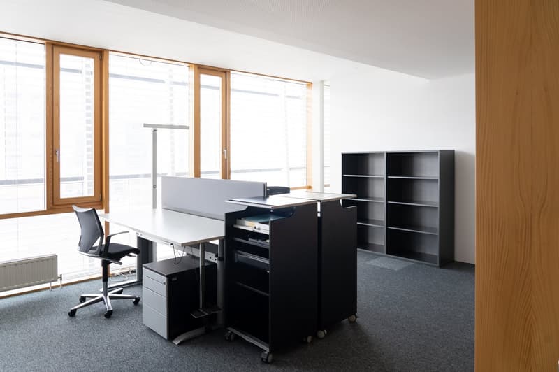 The image shows a modern office space with large windows, a white desk, a black office chair, and several black storage cabinets on wheels. The room has a minimalist and clean design with wooden accents.