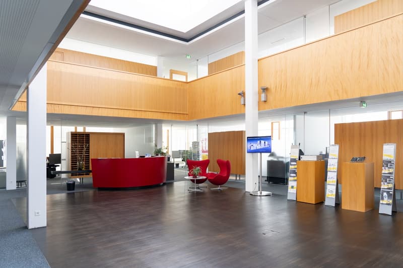 Spacious office lobby with high ceilings, wooden paneled walls, red leather chairs, and a reception desk. There are also display racks and a TV screen visible.