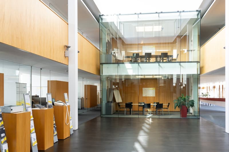 Spacious lobby with glass walls, wooden paneling, and modern furniture. There is a reception desk, chairs, and a conference room visible through the glass walls.