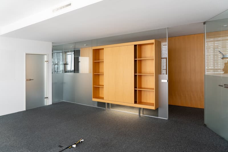 The image shows a modern office space with a large wooden cabinet or shelving unit along one wall. The room has a gray carpeted floor, white walls, and glass partitions. There is a door visible on the left side of the image.