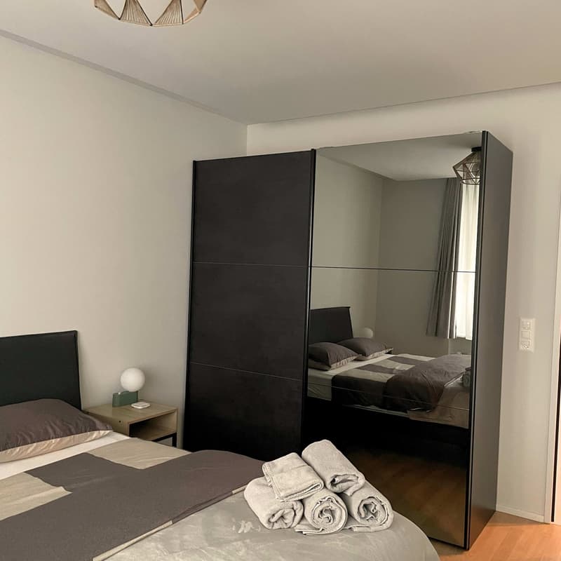 A bedroom with a large mirror and a window behind the bed, a side table with a lamp, a large mirror cabinet, a double bed with pillows and blankets, a nightstand with a lamp, and a ceiling light