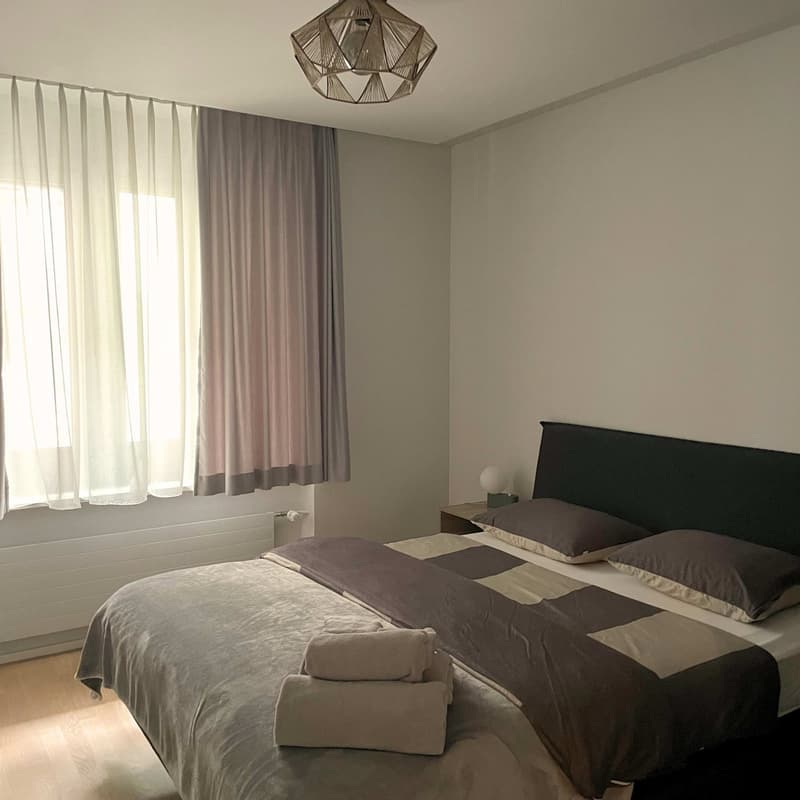 a bed with blanket, pillow and towels, two nightstands with lamp, white and gray curtains
