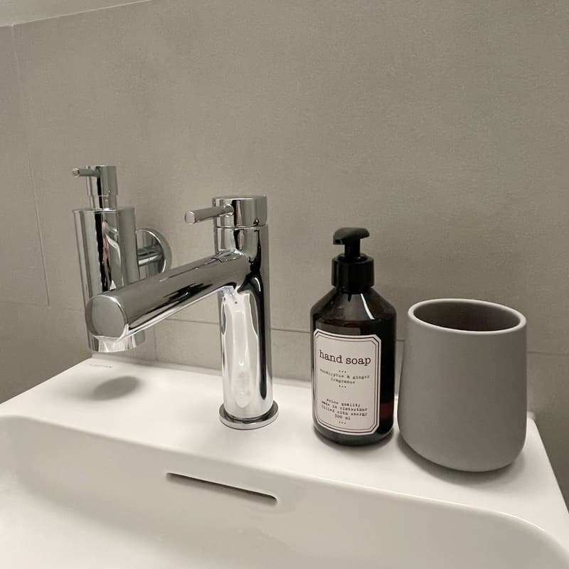 a sink with a faucet, a bottle of hand soap, a white cup, a gray tile wall