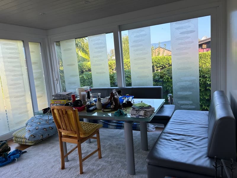 Sunroom with large windows, glass table, yellow chair, leather sofa, white rug, storage boxes, books, and toys