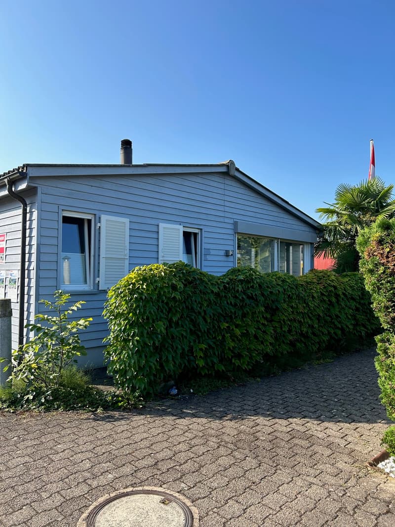 Small detached house with a brick driveway, gray exterior with white shutters, hedges and a small garden in the front