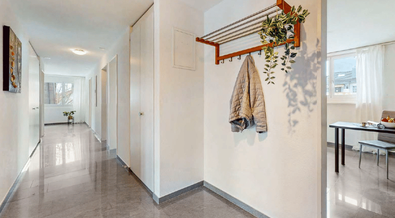 Bright and spacious hallway with tiled flooring, white walls, and a coat rack with a quilted jacket hanging on it. There is a potted plant and a decorative element on the wall. The hallway leads to other rooms in the apartment.