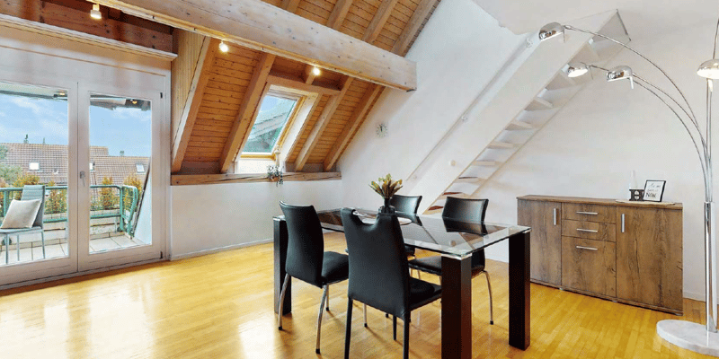 Spacious attic room with wooden beams, large windows, glass dining table, black leather chairs, wooden cabinet, and access to a balcony or terrace.