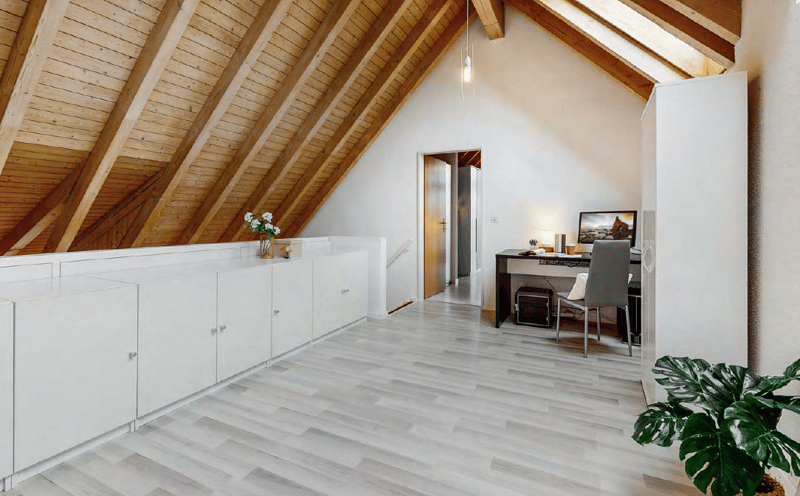 Attic bedroom with wooden beamed ceiling, white walls, hardwood flooring, built-in storage cabinets, desk area, and a view through the window.