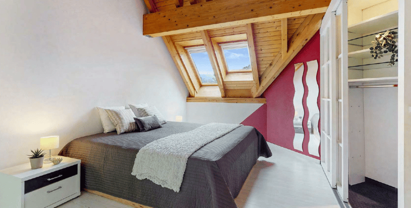 Attic bedroom with wooden beams, slanted ceiling, large windows, gray bedding, and a red accent wall