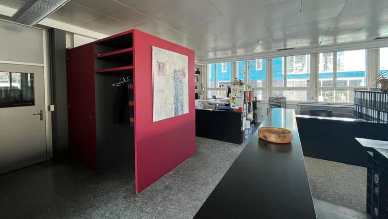 The image shows an office space with a modern and minimalist design. The room has large windows that provide natural light, and the walls are painted in a neutral color scheme. There is a red storage unit or cabinet in the center of the room, and a wooden