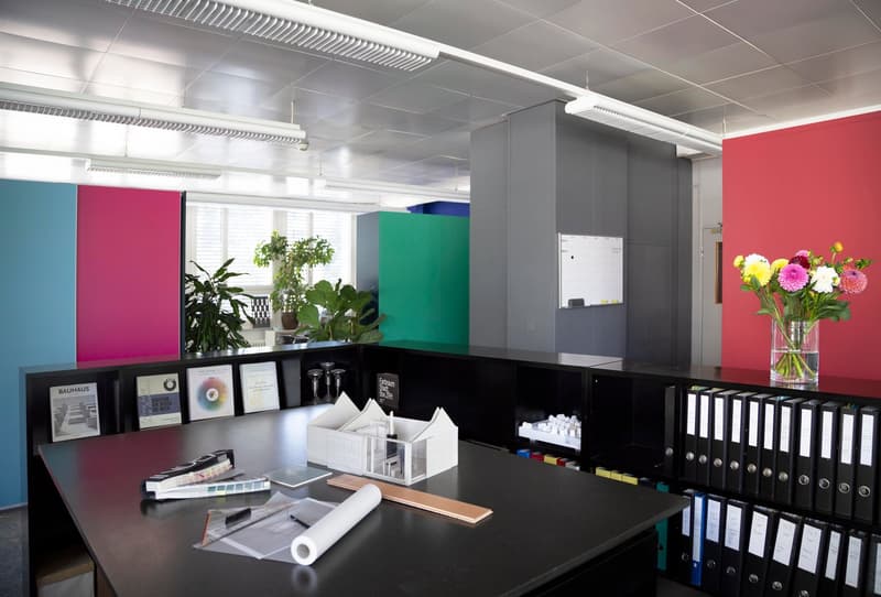 Modern office space with colorful walls, black desks, shelves with office supplies, and a vase of flowers on the desk