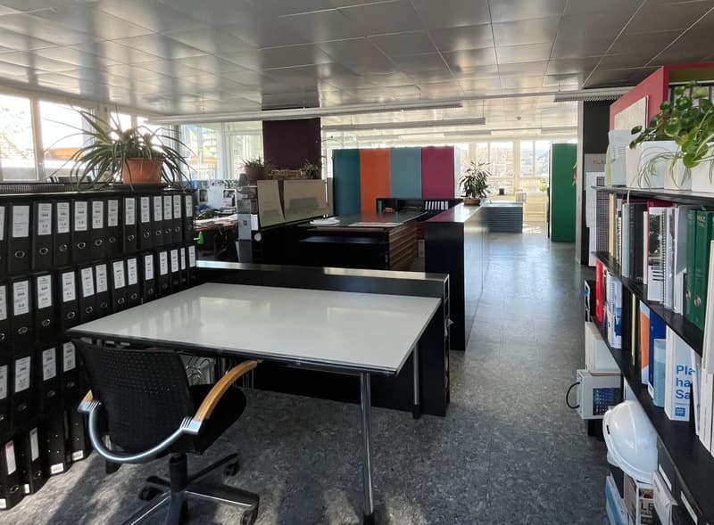 The image shows an open office space with a modern and colorful design. There are rows of black binders on shelves, a large white table in the center, and various office equipment and plants throughout the room. The walls feature colorful panels in differ