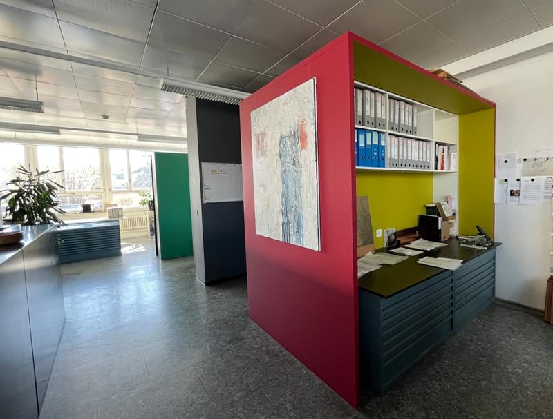 The image shows an office space with a modern, colorful design. There is a red and yellow partition wall with an artwork displayed on it. The office has a reception desk with drawers and shelves for storage. The space also includes a green wall, a plant,
