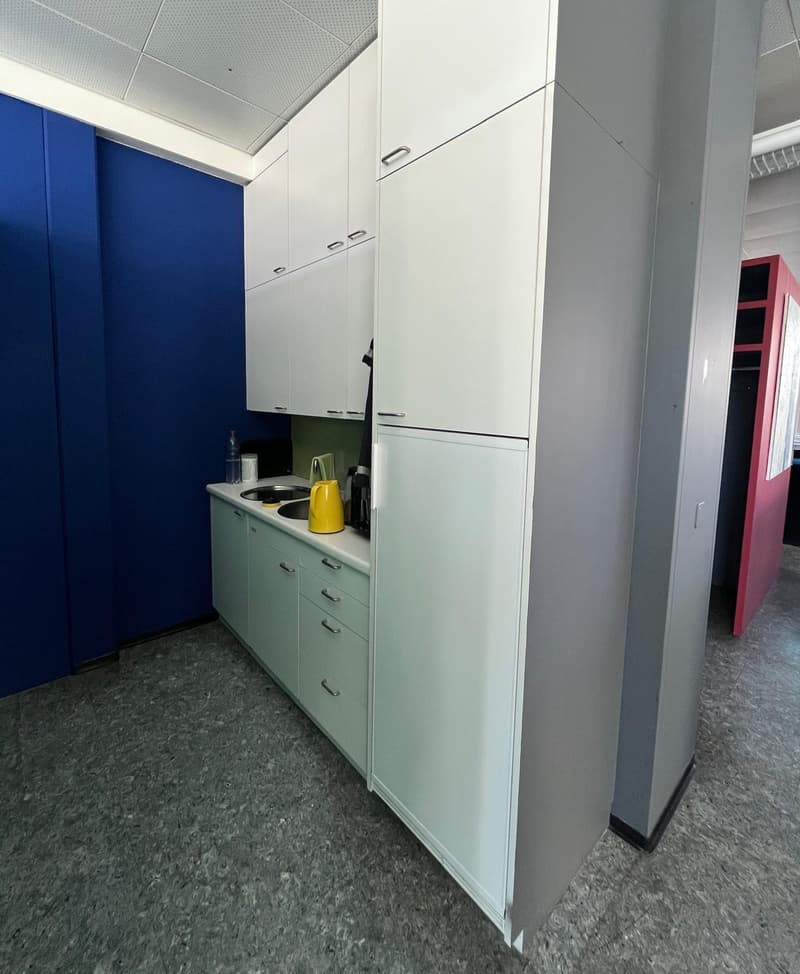 The image shows a small kitchenette area with white cabinets, a sink, and a refrigerator. The floor is a gray tile or linoleum material. The walls are painted in a blue color on one side and a gray color on the other side.