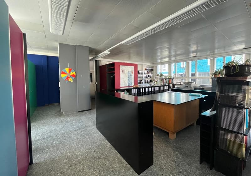 Open office space with colorful accents, including a large spinning wheel decoration. The space features a central desk area, bookshelves, and various office equipment and furnishings.