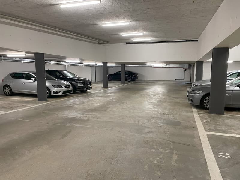 underground car park, concrete floor, illuminated by overhead lights, several cars parked, grey and black colored vehicles