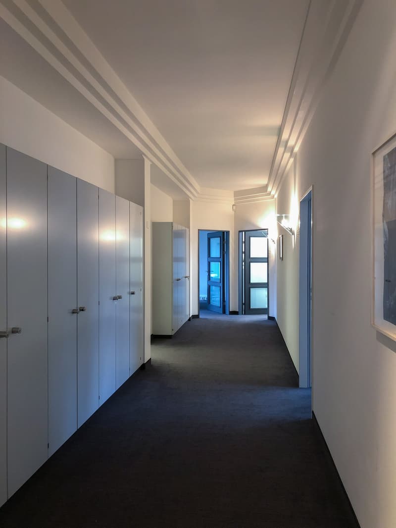 Long hallway with gray built-in cabinets/lockers along one wall, dark carpeted floor, and recessed lighting in the ceiling.
