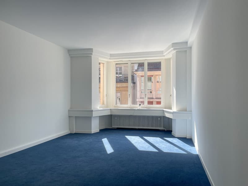 Spacious room with large windows, white walls, and blue carpeted floor. The room features built-in shelving and a radiator along the wall.