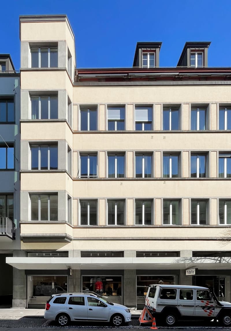 Multi-story commercial building with large windows, balconies, and a parking area in front with a parked car and a utility vehicle